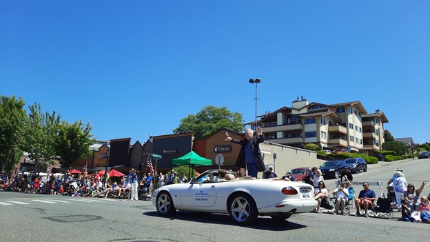 Rick Steves standing in a white corvette during a parade in the summer. 